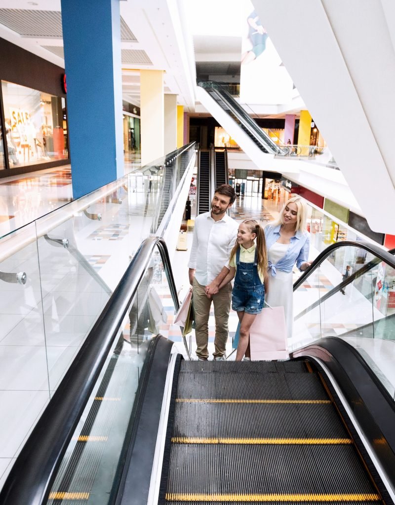Family On Shopping Moving Up Standing On Escalator Stairs In Hypermarket Holding Shopper Bags. Empty Space